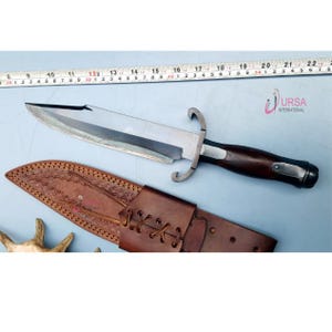 May include: A large, silver-bladed knife with a dark wooden handle and a brown leather sheath. The knife has a curved guard and a serrated edge. The sheath is intricately stitched. The image also includes a measuring tape. The text "URSA INTERNATIONAL" is visible.