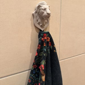 Könnte beinhalten: Ein Wandhaken mit einer skulptierten, hellen Löwenkopf-Plakette. Ein dunkles Handtuch mit einem lebendigen Blumenmuster in Rot-, Orange- und Grüntönen hängt an dem Haken und zeigt einen detaillierten botanischen Druck auf dem Stoff.
