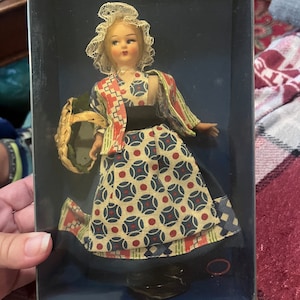 May include: A small porcelain doll wearing a blue, red, and white patterned dress with a white lace bonnet and holding a wicker basket.
