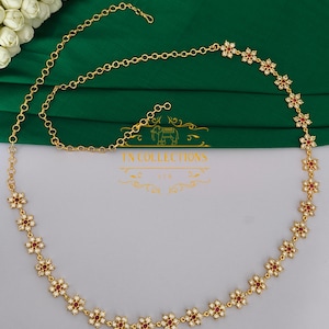 May include: A gold-toned waist chain featuring a series of repeating flower-shaped links. Each flower is accented with small white stones and a central red stone. The chain includes a clasp and an adjustable length section with a logo reading TN COLLECTIONS.