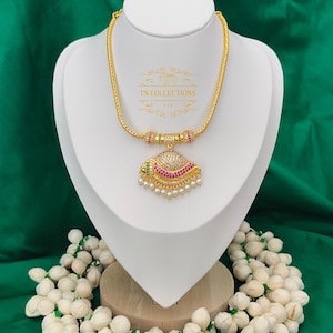 May include: Gold necklace with a pendant adorned with red and white gemstones and pearls. The necklace is displayed on a white bust against a green background. The text "TN COLLECTIONS" is visible on the bust.