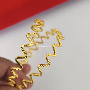 May include: Two gold-coloured, zigzag-patterned bangles. The bangles have a textured surface and are held against a light background. The text "TN Collections" is visible on the bangles.