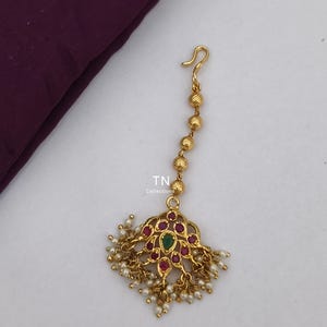 Puede incluir: Tocado dorado con un diseño floral central, adornado con piedras preciosas rojas y verdes y pequeñas perlas blancas. El tocado tiene una cadena de cuentas doradas y un gancho. El texto "TN Collections" es visible.