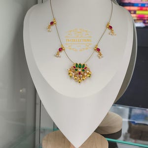 Puede incluir: Collar dorado con un colgante central con piedras preciosas verdes y rojas, rodeado de perlas y detalles dorados. El collar tiene delicados hilos con detalles rojos y blancos. El texto "TN COLLECTIONS" es visible.
