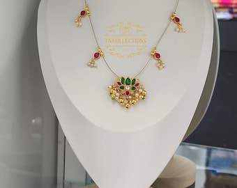 May include: Gold-toned necklace with a central pendant featuring green and red gemstones, surrounded by smaller pearls and gold accents. The necklace has delicate strands with red and white accents. The text "TN COLLECTIONS" is visible.