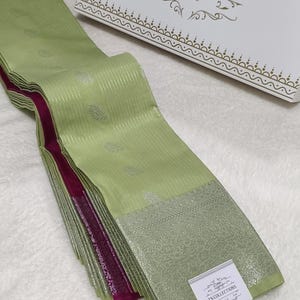 May include: A folded light green saree featuring silver vertical stripes and leaf motifs, with a wide silver border and a deep magenta inner edge. A white tag with the text TN COLLECTIONS is attached to the fabric, near a matching white box.