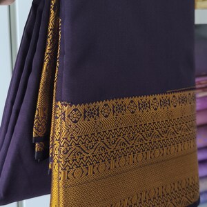 May include: A deep purple fabric textile featuring a wide, ornate border with intricate gold-coloured geometric and floral patterns. The fabric is folded in layers, showcasing the contrast between the dark purple base and the metallic gold detailing.