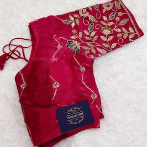 May include: A magenta-colored blouse with intricate gold floral embroidery on the sleeves and neckline. The blouse features a V-neck design and delicate gold embellishments. A small blue tag with the text "TN COLLECTIONS" is visible.
