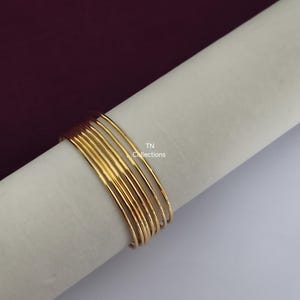 Puede incluir: Un conjunto de pulseras finas de color dorado. Las pulseras están colocadas sobre una superficie blanca, con un fondo burdeos oscuro. El texto "TN Collections" es visible.