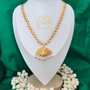 Può includere: Collana dorata con pendente con pietre rosse e bianche, esposta su un manichino bianco. Il pendente presenta un disegno di uccello e piccole perle bianche. Lo sfondo è verde. Collezione TN.