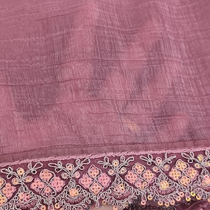 May include: Close-up of a mauve fabric with a textured surface. The bottom edge features an embroidered border with floral and sequin details in shades of pink, orange, and silver. The fabric appears to be lightweight and flowing.