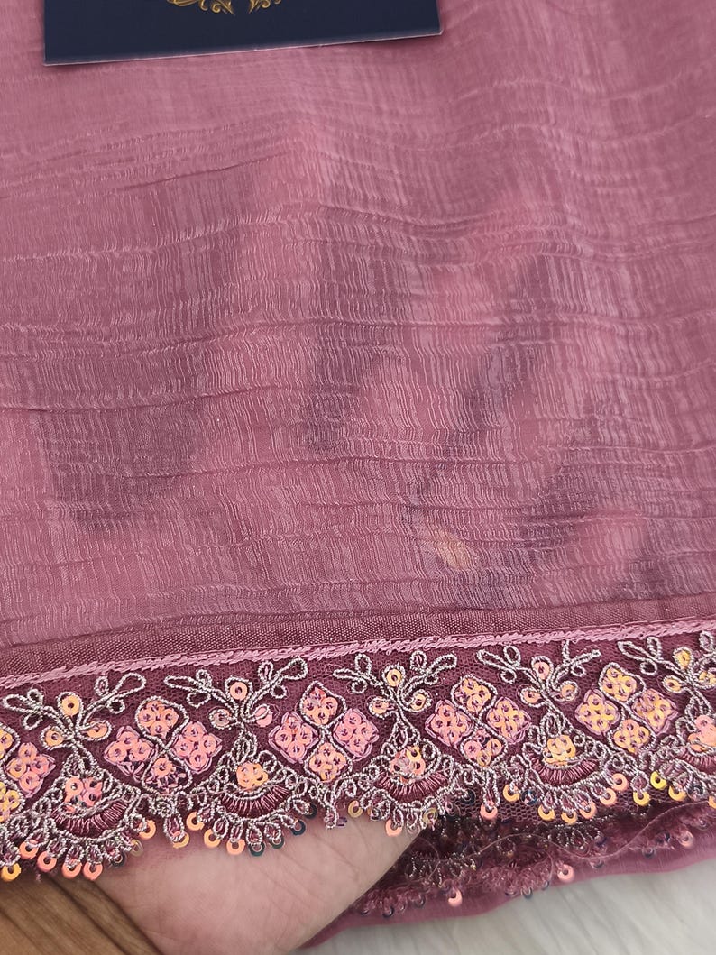 May include: Close-up of a mauve fabric with a textured surface. The bottom edge features an embroidered border with floral and sequin details in shades of pink, orange, and silver. The fabric appears to be lightweight and flowing.