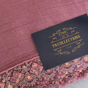 May include: Dusty rose fabric with a detailed, sequined border. The border features intricate floral embroidery and small, shimmering sequins. A navy blue card with gold text that reads "TN COLLECTIONS LTD" is on top of the fabric.