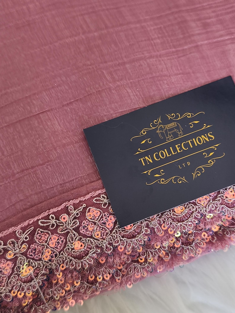 May include: Dusty rose fabric with a detailed, sequined border. The border features intricate floral embroidery and small, shimmering sequins. A navy blue card with gold text that reads "TN COLLECTIONS LTD" is on top of the fabric.