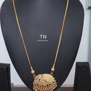 Puede incluir: Collar dorado con un colgante con diseño de pavo real adornado con pequeñas cuentas blancas. El collar se exhibe en un expositor de joyería negro. El texto "TN Collections" es visible.