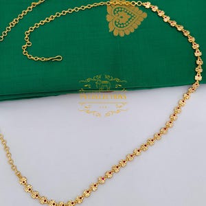 May include: Gold-coloured body chain featuring a floral pattern with small red accents. The chain is displayed on green fabric. The chain has a hook closure and is from TN Collections LTD.