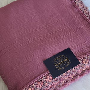 May include: A dusty rose-colored fabric with a textured surface and a decorative border. The border is embellished with sequins and intricate embroidery. A navy blue tag with gold text reads "TN COLLECTIONS LTD."
