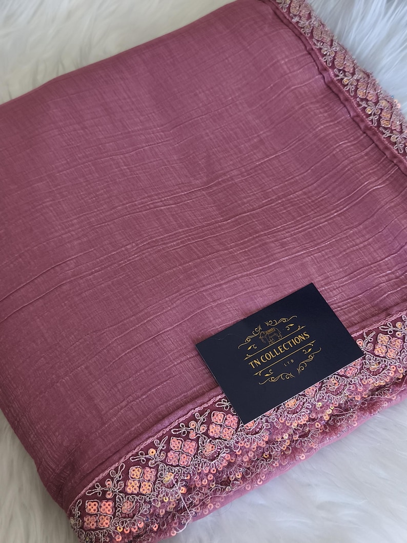 May include: A dusty rose-colored fabric with a textured surface and a decorative border. The border is embellished with sequins and intricate embroidery. A navy blue tag with gold text reads "TN COLLECTIONS LTD."