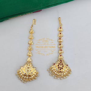 May include: Two gold-colored headpieces with intricate designs. Each piece features a fan-shaped base adorned with white and red gemstones, and small white pearls. The text "TN COLLECTIONS LTD" is visible.