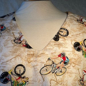 May include: A beige fabric with a repeating pattern of cyclists riding bicycles on a map. The map includes the names of cities and towns in France, including Paris, Vendée, and Olonne sur Mer.