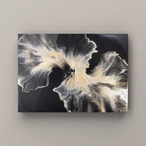 Pode incluir: Obra de arte abstrata com fundo preto e um design floral central em branco, creme e prata. A obra tem um acabamento brilhante e é montada em uma moldura retangular. O design assemelha-se a uma flor desabrochando.