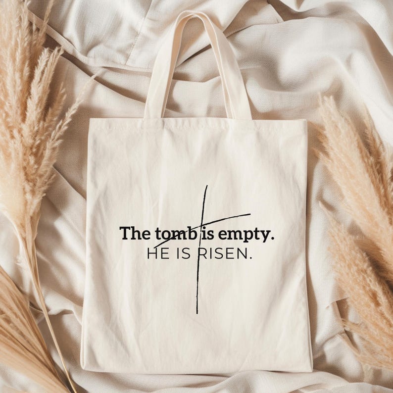 May include: A white canvas tote bag with black text that reads "The tomb is empty. HE IS RISEN." The text is centered on the bag and is surrounded by a simple black cross.