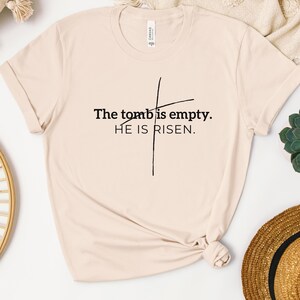 May include: A beige t-shirt with a black cross and the text "The tomb is empty. HE IS RISEN."