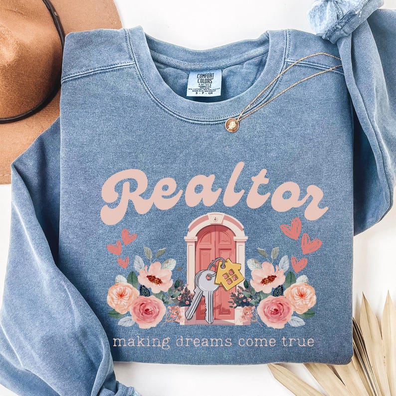 May include: A blue sweatshirt with the text "Realtor making dreams come true" in pink and white lettering. The sweatshirt features a pink door with a key and a house key chain, surrounded by pink flowers.