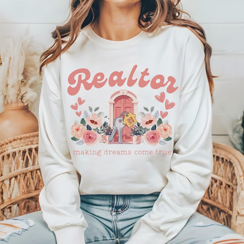 May include: White sweatshirt with a pink graphic design that says "Realtor making dreams come true". The graphic features a pink door with a key and a house key tag, surrounded by pink flowers.
