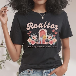 May include: Black t-shirt with the text "Realtor making dreams come true" in pink lettering. The design features a pink door with a key and a house key tag, surrounded by pink flowers.
