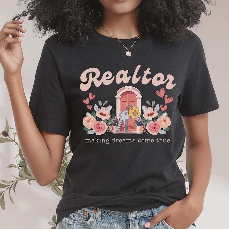 May include: Black t-shirt with the text "Realtor making dreams come true" in pink lettering. The design features a pink door with a key and a house key tag, surrounded by pink flowers.
