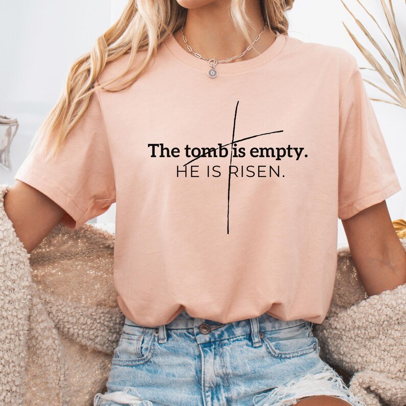 May include: A peach-colored t-shirt with a black cross and the text "The tomb is empty. HE IS RISEN."