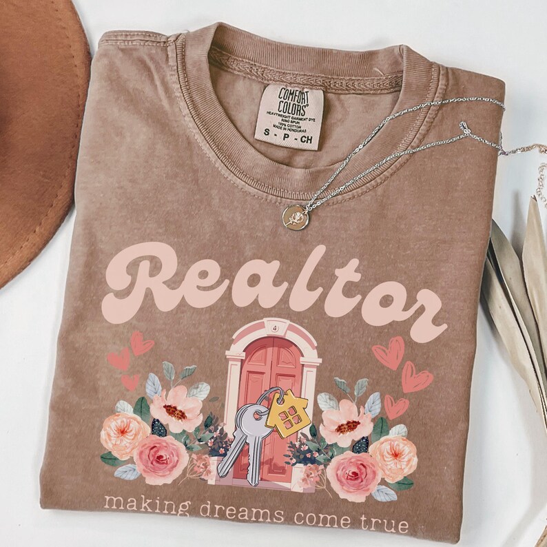 May include: A brown t-shirt with the text "Realtor making dreams come true" in pink script. The design features a pink door with keys and floral accents.