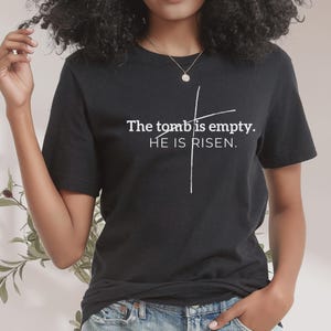 May include: Black t-shirt with white text that reads "The tomb is empty. HE IS RISEN." A white cross is drawn behind the text.