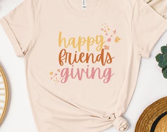 May include: A beige t-shirt with the text "happy friends giving" in orange, yellow, and pink lettering. The text is surrounded by small orange, yellow, and pink leaf designs.