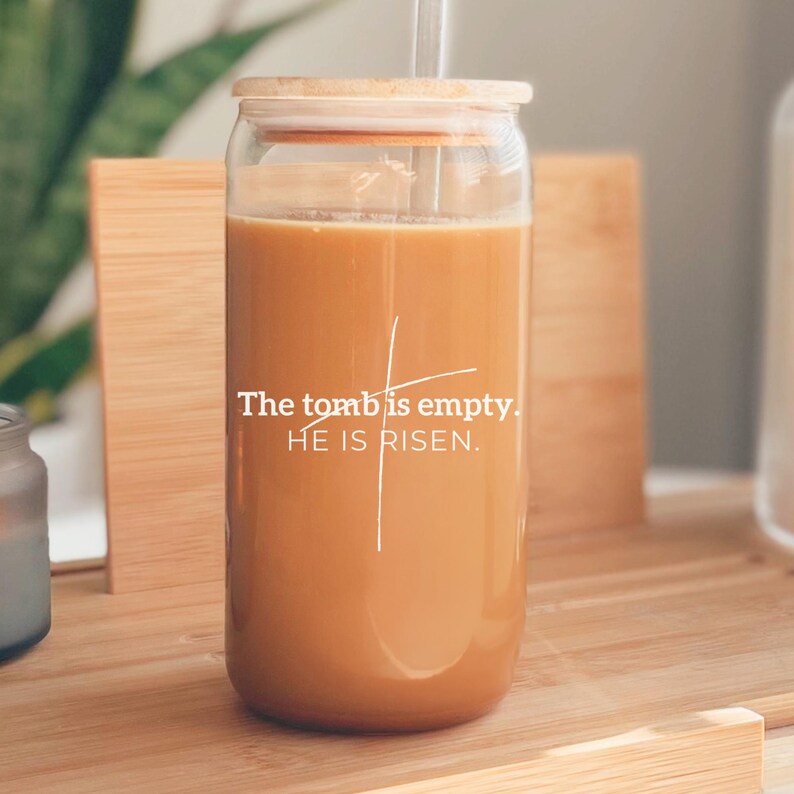 May include: A glass jar with a wooden lid and a straw, filled with a light brown liquid. The jar has a white cross and the text "The tomb is empty. HE IS RISEN." printed on it.