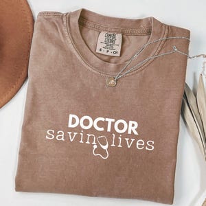 May include: A brown t-shirt with the text "DOCTOR saving lives" in white lettering and a white stethoscope graphic.