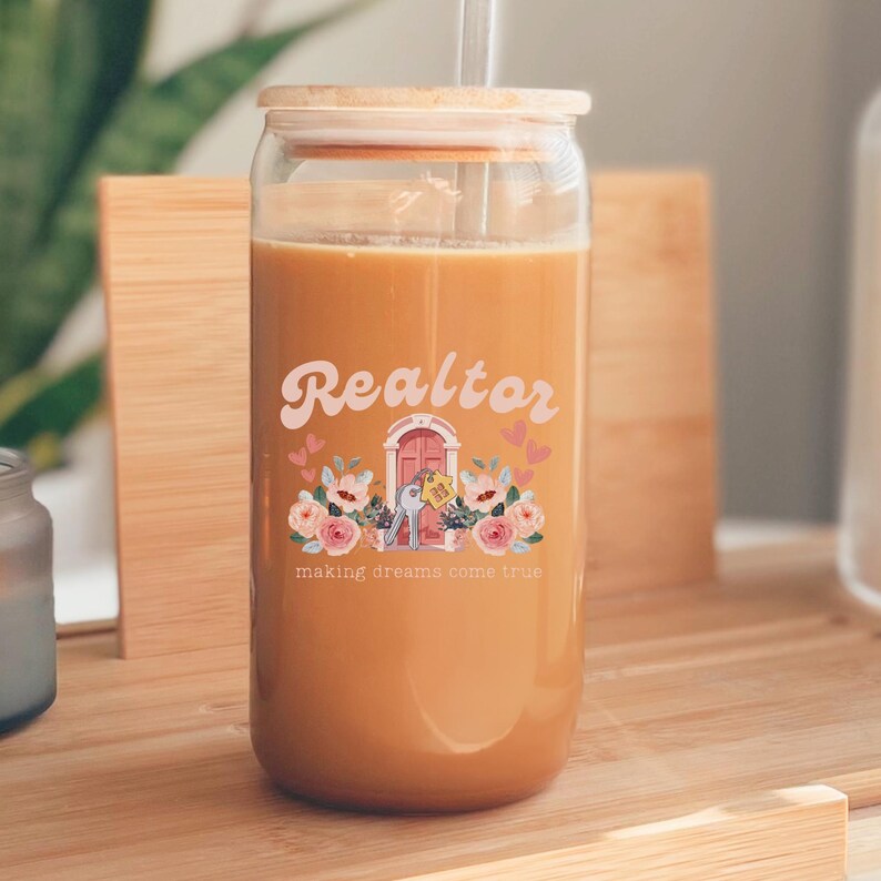 May include: A glass tumbler with a brown lid and a straw. The tumbler has a floral design with a pink house and the text "Realtor making dreams come true".