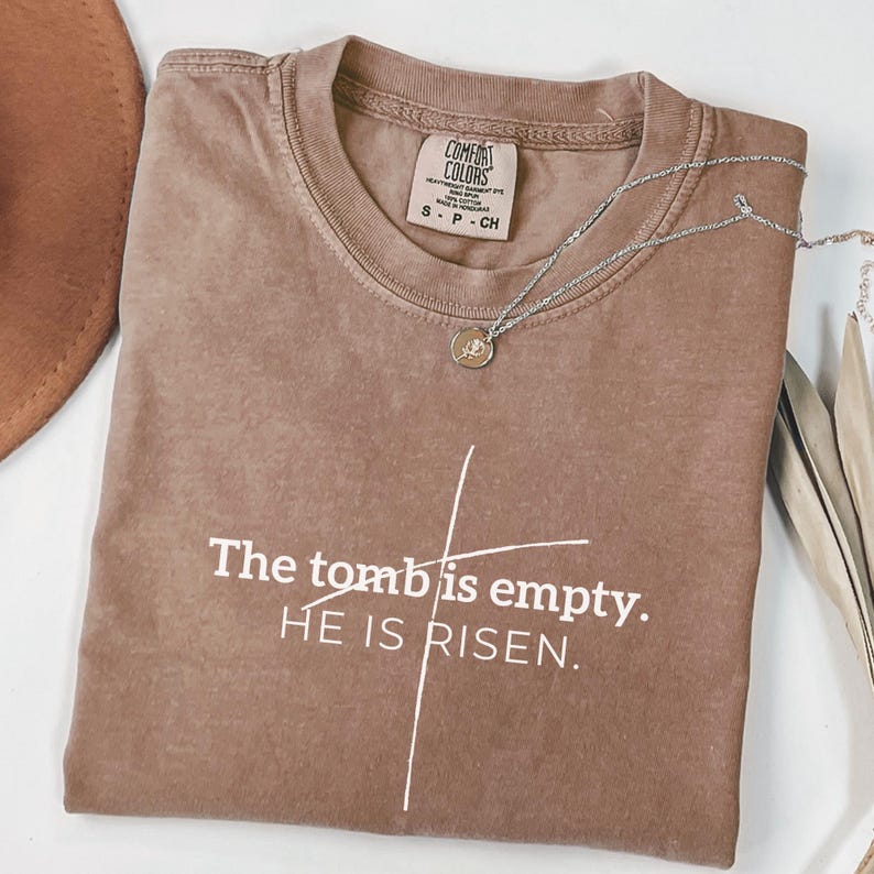 May include: A brown t-shirt with the text "The tomb is empty. HE IS RISEN." printed in white. A simple cross is drawn above the text.
