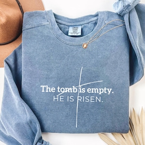 May include: A blue sweatshirt with the text "The tomb is empty. HE IS RISEN." in white lettering. The sweatshirt has a distressed look and a white cross graphic.