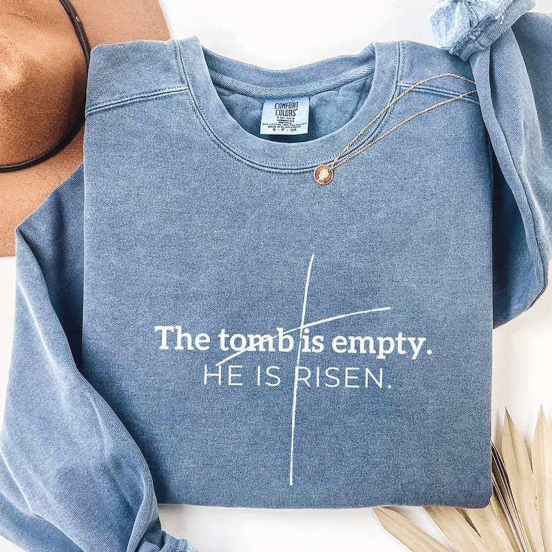 May include: A blue sweatshirt with the text "The tomb is empty. HE IS RISEN." in white lettering. The sweatshirt has a distressed look and a white cross graphic.