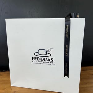 May include: White box with a black ribbon and the text "Fedoras by Gentleman" and the website address "www.fedorasbygentleman.com".