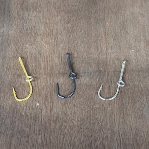 May include: Three fishing hooks in gold, black, and silver are arranged on a wooden surface. Each hook has a curved point and a looped eye for attaching to a fishing line. The hooks are designed for catching fish.
