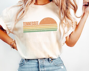 Könnte beinhalten: Ein elfenbeinfarbenes Kurzarm-T-Shirt mit einem Grafikdruck, der das Wort TENNESSEE in orangefarbenen Buchstaben über einer Halbkreis-Sonne und drei horizontalen Streifen in Orange, Gelb und Grün zeigt, alles mit einer abgenutzten Vintage-Textur.