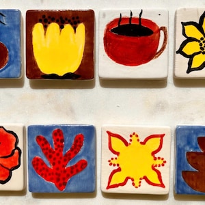 Puede incluir: Un conjunto de ocho baldosas de cerámica cuadradas con diseños pintados a mano. Las baldosas presentan varias ilustraciones coloridas, incluyendo flores, una taza de café y formas abstractas. Cada baldosa mide aproximadamente 5 cm cuadrados.