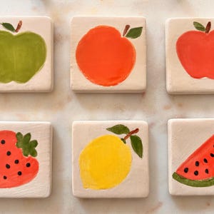 Puede incluir: Seis baldosas de cerámica cuadradas con diseños de frutas pintados a mano. Las frutas incluyen una manzana verde, una manzana naranja, una manzana roja, una fresa, un limón y una rodaja de sandía. Cada baldosa mide aproximadamente 7,6 cm cuadrados.