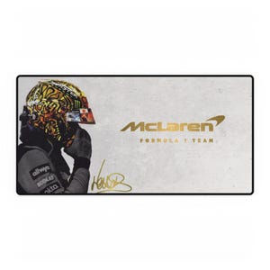 Op de afbeelding: Een rechthoekige muismat met een Formule 1-coureur in een goud-zwarte helm. Het McLaren-logo en de tekst zijn goudkleurig, evenals een gouden handtekening. De achtergrond is lichtgrijs.