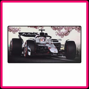 May include: A white Formula One racing car with red and black accents drives on a track with pink cherry blossom trees in the background.