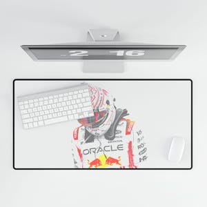 May include: A rectangular desk mat featuring a graphic of a race car driver wearing a white helmet and a white racing suit with red accents, the Oracle logo, and the Red Bull logo. The mat is positioned under a white computer keyboard and mouse.