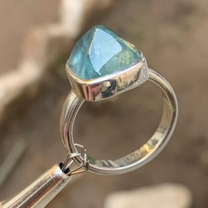 May include: A silver ring featuring a triangular blue gemstone. The ring has a simple band and the gemstone is faceted.